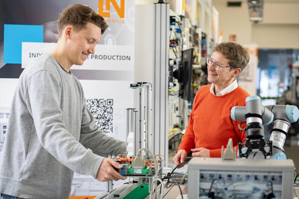 Ein Mann und eine Frau arbeiten an einer Industrieanlage mit einem Roboterarm und Steuerungskomponenten, einer davon lächelt.