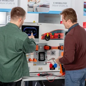 Zwei Männer betrachten und interagieren mit einem Schulungssystem für E-Mobilität, das ein Elektrofahrzeug-Diagramm und Komponenten zeigt.