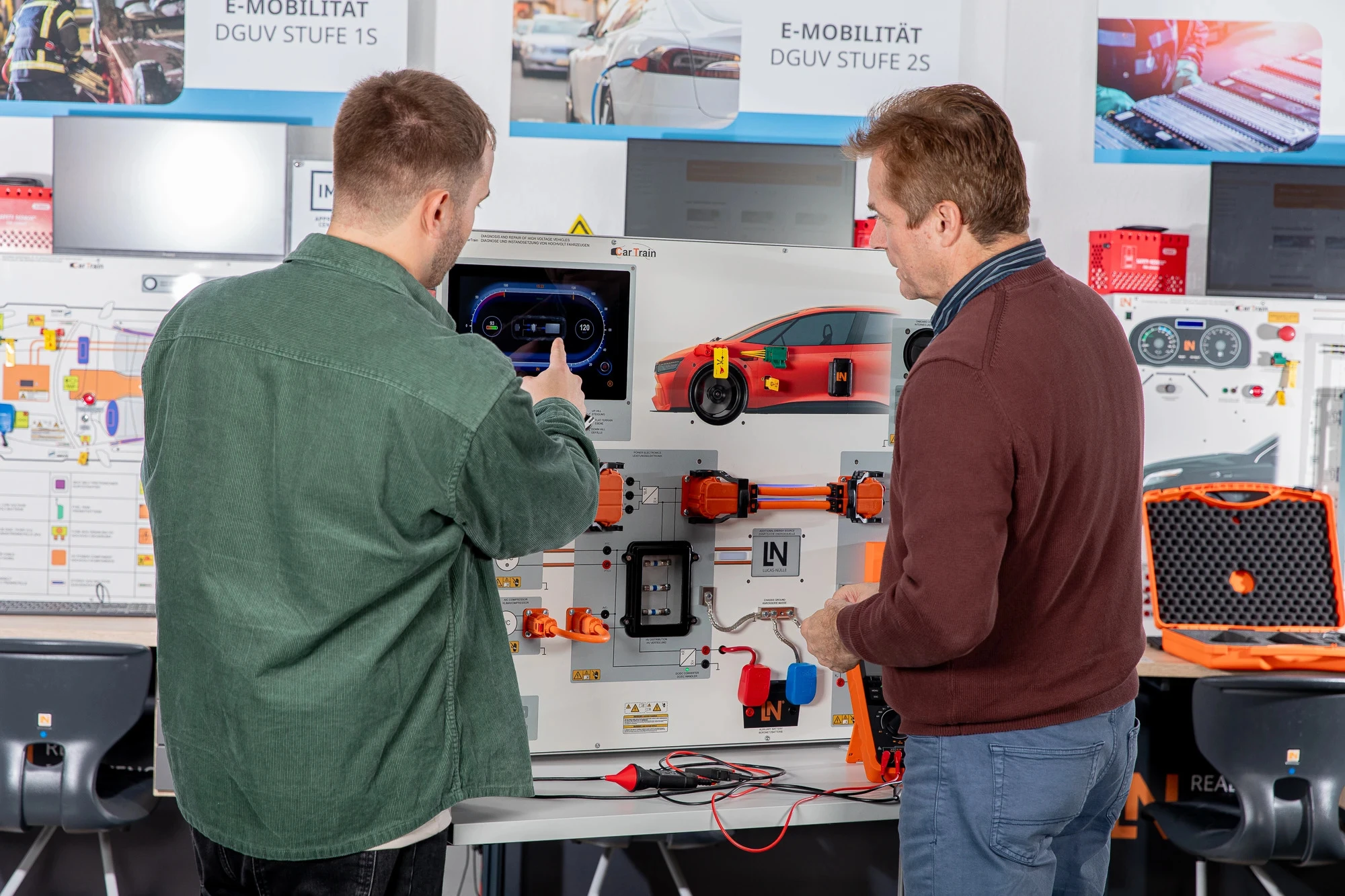 Zwei Männer betrachten und interagieren mit einem Schulungssystem für E-Mobilität, das ein Elektrofahrzeug-Diagramm und Komponenten zeigt.